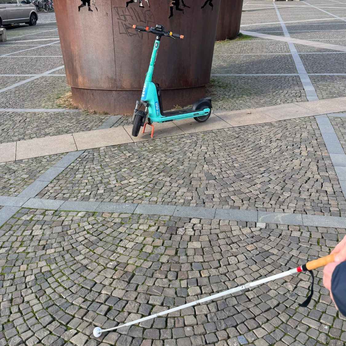 Typische Hindernisse für eine blinde oder sehbehinderte Person im öffentlichen Raum Beim überqueren eines Platzes stehen Kunstwerke und ein Scooter im Weg und erschweren den Weg zum Ziel